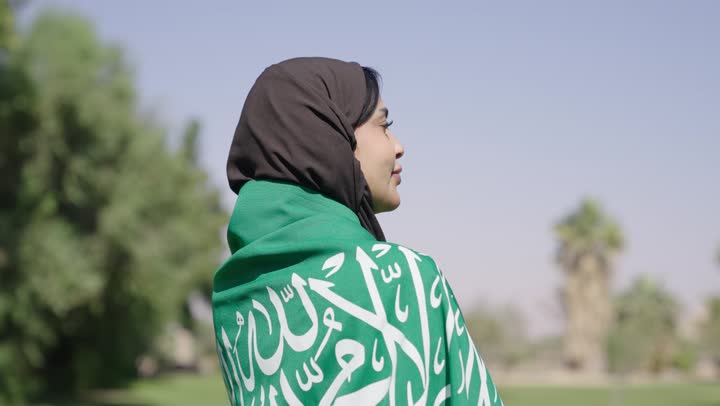 A Saudi Arabian Gulf woman wearing a black abaya and draped in a green flag, with the Saudi flag carried over her shoulders as a symbol of love, pride, and honor.