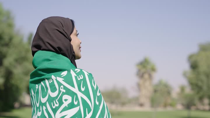 رائحة النسيم العليل و الزهور المتفتحة تملأ الحديقة , امرأة عربية خليجية سعودية محجبة ترتدي عباءة سوداء تتوشح بالعلم السعودي الاخضر , مفهوم حب الوطن