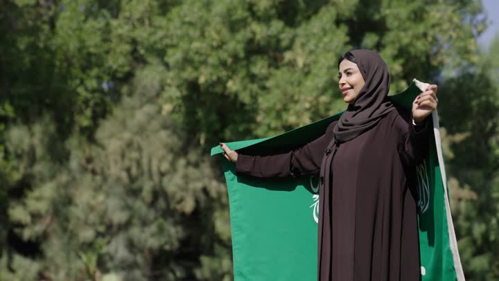 الشعور بالاعتزاز و الفخر اثناء حمل العلم السعودي , راية التوحيد الخضراء ترفرف في الهواء الطلق وسط الطبيعة الساحرة , امرأة عربية خليجية سعودية محجبة ترتدي العباءة السوداء