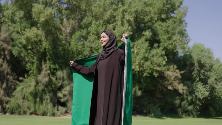 امراة عربية خليجية سعودية محجبة ترتدي العباءة السوداء ,الشعور بالسعادة و الفخر اثناء حمل العلم السعودي , مفهوم حب الوطن