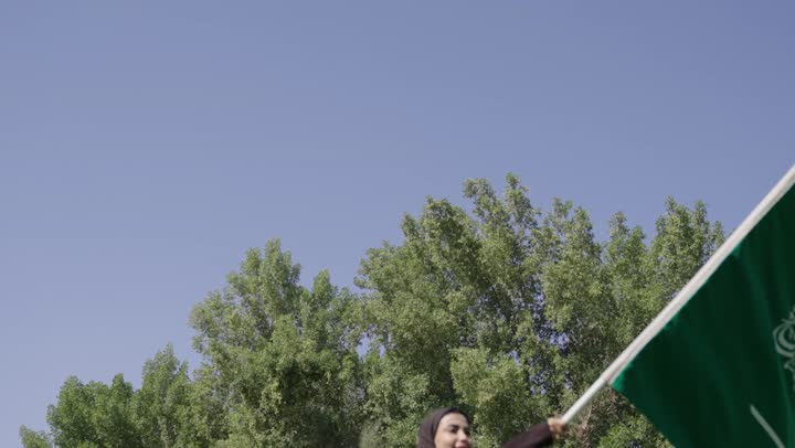 ايماءات السعادة و الفرحة , امراة عربية خليجية سعودية محجبة ترتدي عباءة سوداء تحمل العلم السعودي , الراية الخضراء تطير في الهواء المنعش ,