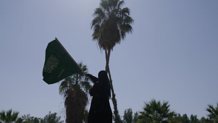 العلم السعودي يحلق عالياً في السماء الصافية , امرأة عربية خليجية سعودية محجبة ترتدي عباءة سوداء تلوح بالراية الخضراء , مساحات خضراء واسعة , جو منعش جميل