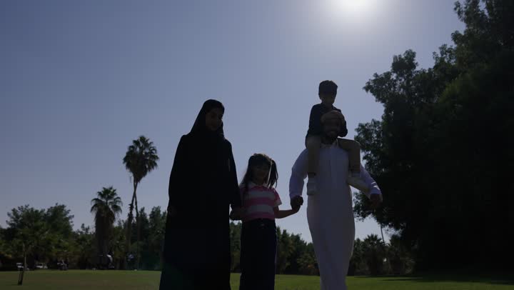 The concept of a happy family, a Saudi Arabian Gulf man wearing traditional attire carries his son on his shoulders, a young boy enjoying playing with his father, a Saudi Arabian Gulf girl dressed in casual wear holding her parents' hands and walking lightly and playfully, a Saudi Arabian woman in a hijab wearing a black abaya enjoying sharing happy moments with her family.