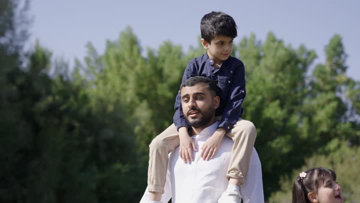 حديث شيق بين الاب و ابنه اثناء التجول في الحديقة , رجل عربي خليجي سعودي يرتدي الثوب التقليدي الابيض يحمل ابنه على الاكتاف , ولد صغير يجري محادثة ممتعة مع والده اثناء المشي بين الاشجار و المساحات الخضراء , مفهوم الترابط الاسري