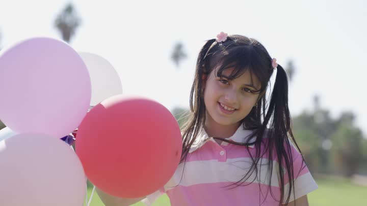 قضاء الاوقات المميزة وسط الجو المشمس والسماء الصافية , فتاة عربية خليجية سعودية صغيرة ترتدي الزي الكاجوال ترفع مجموعة من البالونات الملونة في مرح و سعادة , الابتسامة للكاميرا