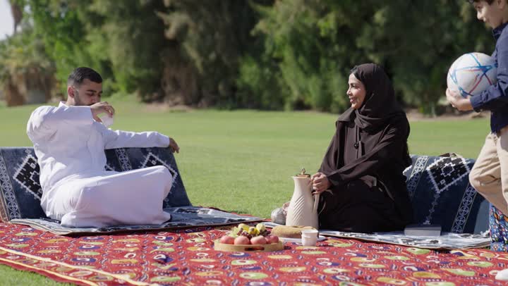 The warm sunny weather and clear sky, a family outing amidst the green spaces and beautiful gardens, an Arab Gulf Saudi man wearing a traditional white thobe warmly welcomes his son, an Arab Gulf Saudi woman in a hijab wearing a black abaya hands the father a cup of warm coffee, a little girl holding a bunch of balloons, unique family moments.