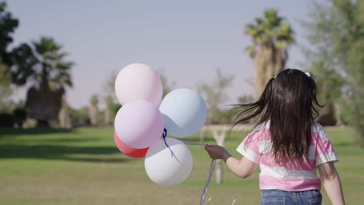 طفلة عربية خليجية سعودية ترتدي الزي الكاجوال تمسك مجموعة من البالون الملون و تجري بخفة و سعادة , الاستمتاع بالهواء الطلق و الطبيعة الجميلة