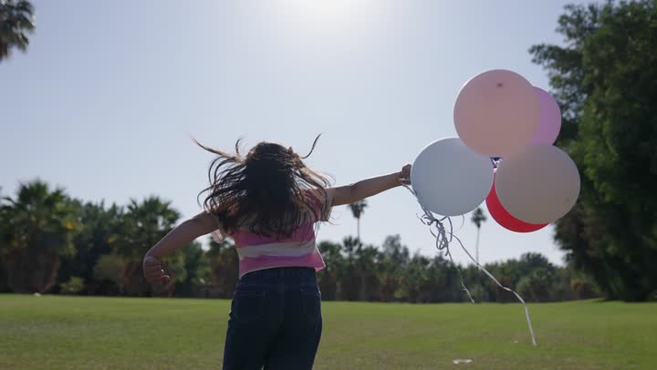 اللعب و اللهو وسط الطبيعة الخلابة والمساحات الخضراء , طفلة عربية خليجية سعودية ترتدي الزي الكاجوال تحمل مجموعة من البالونات الملونة,الاستمتاع بالهواء الطلق و الشمس الساطعة