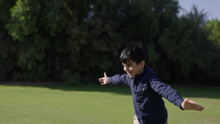Letting children soar in the air, a Gulf Arab Saudi child wearing casual attire spreads his arms as a sign of flying, an Arab Saudi father dressed in traditional white thobe lifts his son into the air among the trees and green spaces, a family outing and unforgettable memories.
