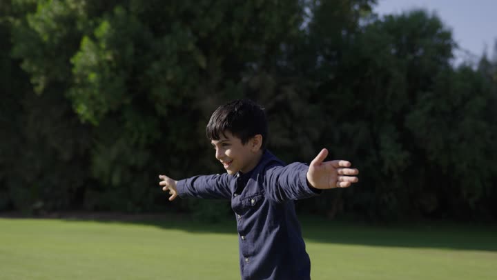 A small Arab Gulf Saudi boy wearing casual attire runs joyfully towards his father, a Gulf Arab Saudi man dressed in traditional white thobe welcomes his son happily. Circular movements in the air indicate flying, a family outing in the warm atmosphere.