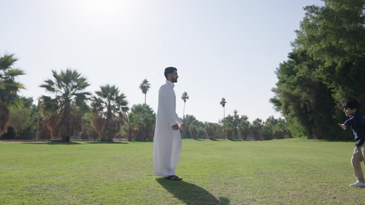 A Saudi Arabian Gulf Arab boy wearing casual attire runs towards his father with happiness and reassurance. The father, a Saudi Gulf Arab dressed in a traditional white thobe, lifts his son joyfully and spins in circles, flying in the open air and enjoying the warm atmosphere.