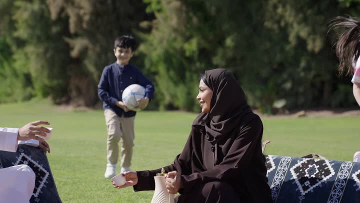 اوقات عائلية سعيدة بين الاشجار و النخيل , اطفال صغار يجرون للاب و الام في مرح طمأنينة ,ام عربية خليجية سعودية محجبة ترتدي عباءة سوداء تجلس بجوار الاب في الحديقة , اب عربي خليجي سعودي يرتدي الثوب التقليدي الابيض يستقبل ابنه بحنان و احتضان , مفهوم النزهة العائلية الجميلة