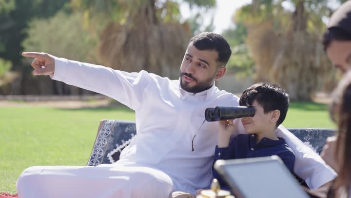 The warm sun and clear sky, an Arab Gulf Saudi father wearing the traditional white Saudi thobe sits next to his son in the garden, an Arab Gulf Saudi boy dressed in casual attire holds a telescope and watches something in the distance, moments of closeness and family bonding.