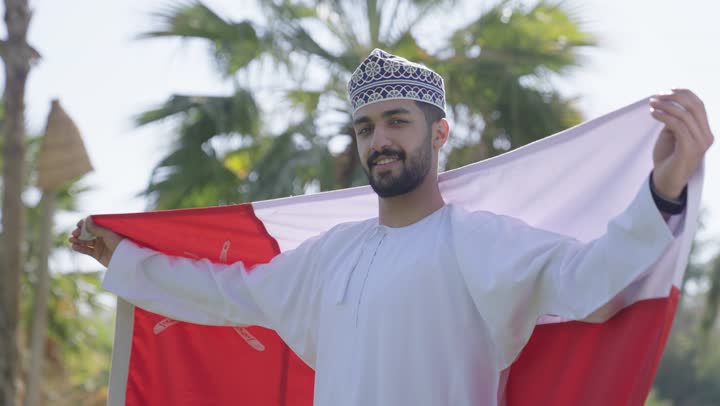 نظرة فخر و اعتزاز بالوطن اثناء حمل العلم العماني , رجل عربي خليجي عماني يرتدي الدشداشة التقليدية و الكمة المطرزة يعقد ذراعيه محتضناً راية الوطن