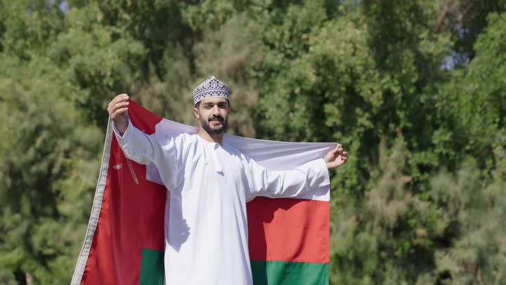 The feeling of warmth under the bright sun and embracing the national flag, an Arab Gulf Omani man wearing the traditional white dishdasha in the Sultanate of Oman crosses his arms while holding the Omani flag, enchanting nature, distinctive atmosphere.