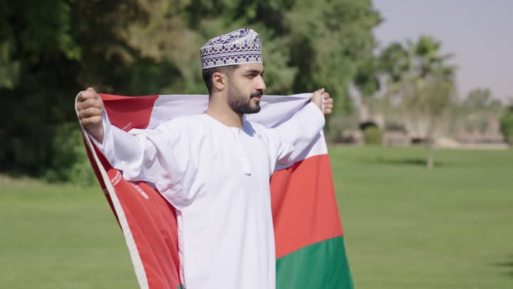 الالتفات للكاميرا مع نظرة الفخر و العزة , شاب عربي خليجي عماني يرتدي الدشداشة البيضاء التقليدية و الكمة المطرزة يضع العلم العماني على ظهره , اوقات ممتعة تحت سماء الوطن