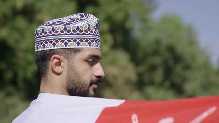Expressions of pride and dignity while raising the Omani flag, a young Arab Gulf Omani man wearing a white dishdasha and a embroidered kumma carries the banner of the Sultanate on his shoulder, the concept of defending the homeland and the safety of its lands.