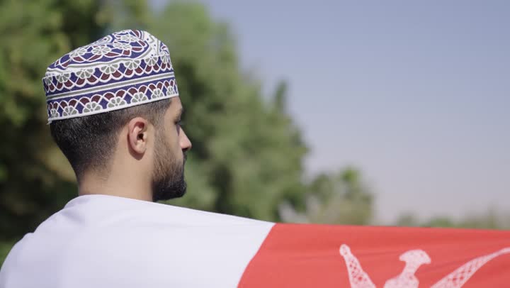 الالتفاف للكاميرا في حركة دائرية , رجل عربي خليجي عماني يرتدي الدشداشة البيضاء و الكمة المطرزة يحمل العلم العماني فوق اكتافه , مفهوم حب الوطن , الاحساس بالامن و الامان تحت سماء الوطن