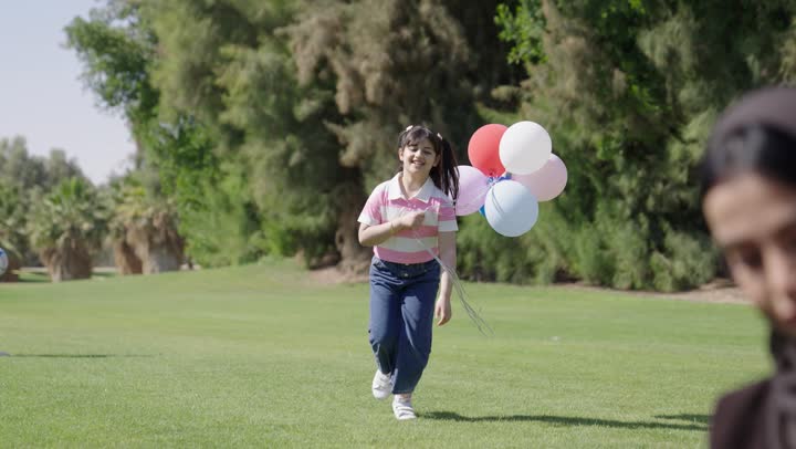 طفلة عربية خليجية عمانية ترتدي الزي الكاجوال تحمل البالونات الملونة في سعادة , الجرب بخفة و رشاقة نحو الام و الاب , سيدة عربية خليجية عمانية محجبة تستقبل الابناء الصغار بفرحة و بهجة , رجل عربي خليجي عماني يرتدي الدشداشة البيضاء و الكمة المطرزة يستمتع بالقهوة العربي الساخنة وسط الاجواء العائلية المميزة