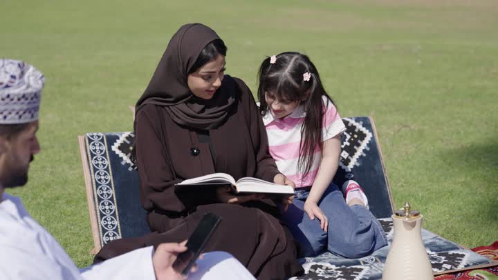 الاستمتاع بقراءة الكتب الشيقة وسط المساحات الخضراء , امرأة عربية خليجية عمانية محجبة ترتدي عباءة سوداء تجلس بجوار الابنة الصغيرة , طفلة عربية خليجية عمانية ترتدي الزي الكاجوال تقرأ الكتاب مع الام بايماءات السعادة والبهجة , رجل عربي خليجي عماني يرتدي الدشداشة البيضاء و الكمة المطرزة يمسك الهاتف المحمول يتابع التطورات في العالم التقني