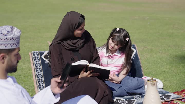 A family gathering between the mother and young children amidst natural scenery, an Omani Gulf Arab mother in a black abaya sits with the family enjoying an interesting book, an Omani Gulf Arab girl in casual attire sits next to her mother listening attentively to the story, an Omani Gulf Arab man wearing a white dishdasha and an embroidered kumma browses his mobile phone.