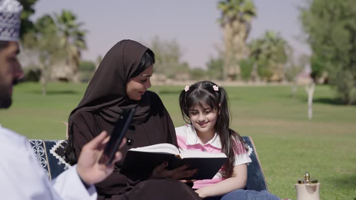الجلسات الخارجية في الهواء الطلق مع استخدام التقنية الحديثة , ام عربية خليجية عمانية محجبة ترتدي عباءة سوداء تروي لابنتها القصص و الحكايات المسلية , طفلة صغيرة تجلس بجوار امها تستمع للقصة بانصات , اب عربي خليجي عماني يجلس بجوار عائلته و يحمل الهاتف المحمول