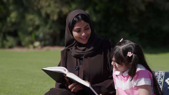 الجلوس في الحديقة و الاستمتاع بانزهة الخارجية في الاجواء الدافئة , اب عربي عماني خليجي يرتدي الدشداشة البيضاء و الكمة المطرزة يحتسي القهوة العربي في رواق و يتصفح الهاتف المحمول , ام عربية خليجية عمانية محجبة ترتدي عباءة سوداء تحكي لابنتها الصغيرة القصص الممتعة و الحكايات التراثية