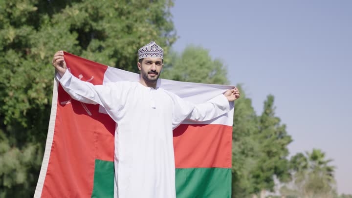 الشعور بالامان داخل الوطن , رجل عربي خليجي عماني يرتدي الدشداشة البيضاء و الكمة المطرز ة يحتن العلم العماني اشارة على الاحساس بالامن و الاطمئنان داخل ارض  الوطن , مناظر طبيعية مبهجة