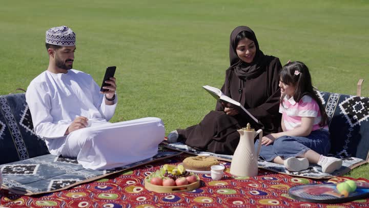 جلسة عائلية في احضان الطبيعة الخلابة , اب عربي خليجي عماني يرتدي الدشداشة البيضاء و الكمة المطرزة يجلس بجوار عائلته و يتصفح الهاتف المحمول , ام عربية خليجية عمانية محجبة ترتدي عباءة سوداء تقرأ مع ابنتها القصص المسلية , قضاء لحظات اسرية ممتعة
