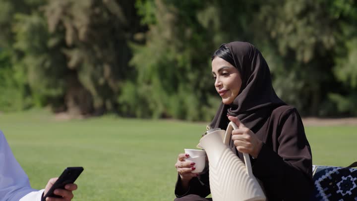 نزهة عائلية ممتعة وسط المناظر الطبيعية الساحرة , رجل عربي خليجي عماني يرتدي الدشداشة البيضاء و الكمة المطرزة يحمل الهاتف المحمول يجلس بجوار زوجته , اراة عربية خليجية عمانية محجبة ترتدي عباءة سوداء تملأ كوب القهوة لزوجها للاستمتاع بالمذاق العربي المميز  , الابنة الصغيرة تجري و تلعب بجوار الوالد و الوالدة بالبالونات في سعادة