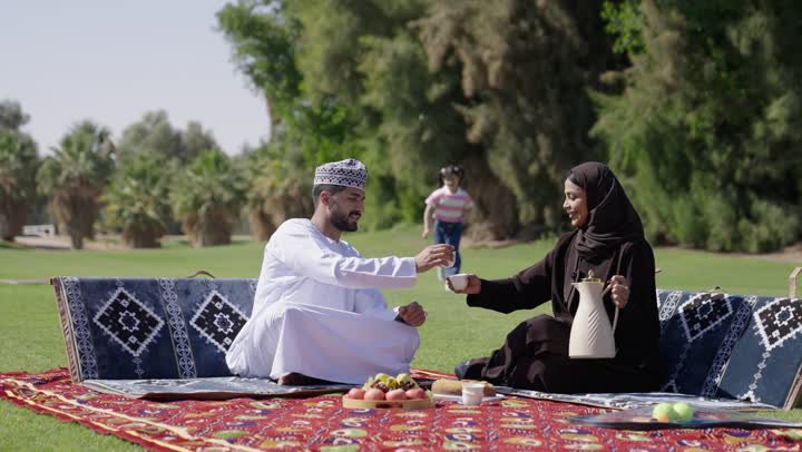 فتاة صغيرة عربية خليجية عمانية ترتدي الزي الكاجوال تجري نحو والدها تغمض عينه و تمازحه , وقت اللعب و اللهو مع العائلة . امرأة عربية خليجية عمانية محجبة ترتدي عباءة سوداء تملأ كوب القهوة العربي لزوجها , رجل عربي خليجي عماني يرتدي الدشداشة البيضاء و الكمة المطرزة يجلس بجوار زوجته , اوقات عائلية مميزة