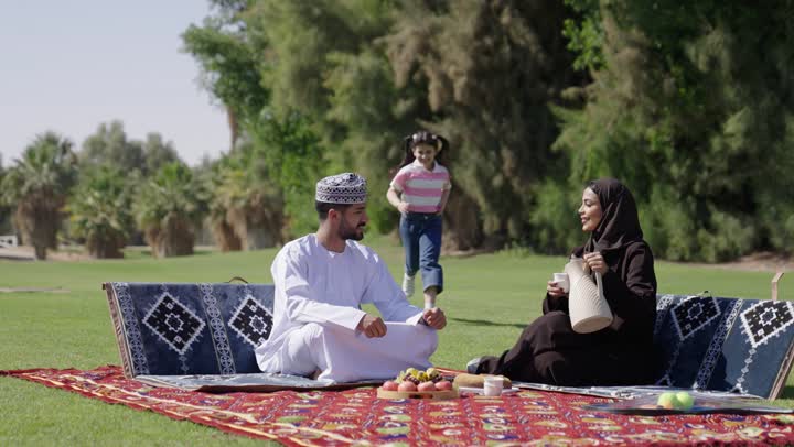 ابنة عربية خليجية عمانية ترتدي الزي الكاجوال تجري نحو والدها تمازحه , رجل عربي خليجي عماني يرتدي الدشداشة البيضاء و الكمة المطرزة يجلس مع الزوجة يستمتع بالجو الدافئو الهواء الطلق , امراة عربية خليجية عمانية محجبة  ترتدي عباءة سوداء تهتم بعائلتها