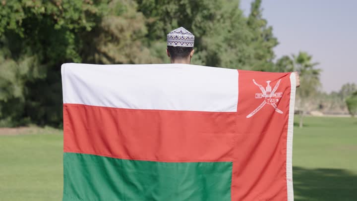 نظرات الشجاعة على وجه الرجل اثناء حمل العلم , رجل عربي خليجي عماني يحمل العلم العماني على ظهره و يعقد ذزاعه محتضناً به, حدائق خضراء واسعة , مناظر طبيعية خلابة