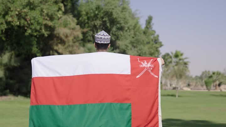 Looking at the camera while wrapping himself in the Omani flag, an Arab Gulf Omani man wearing a white dishdasha and a embroidered kumma carries the Omani flag over his shoulders as a symbol of protection and defense of the homeland.