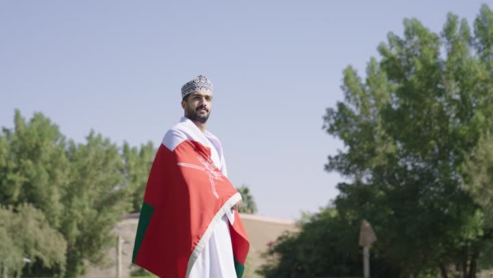 النظر للمستقبل المطمئن داخل الوطن الامن , رجل عربي خليجي عماني يرتدي الدشداشة البيضاء و الكمة المطرزة يضع العلم العماني على ظهره , الجو مشمس و الطبيعة ساحرة