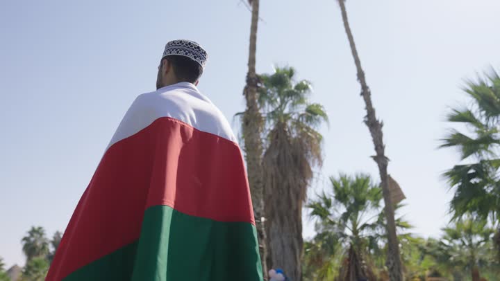 حركة دائرية للكاميرا اثناء التصوير , رجل عربي  خليجي عماني يرتدي الدشداشة البيضاء و الكمة المطرزة , حمل العلم العماني على الاكتاف , الاستمتاع بالاجواء الدافئة