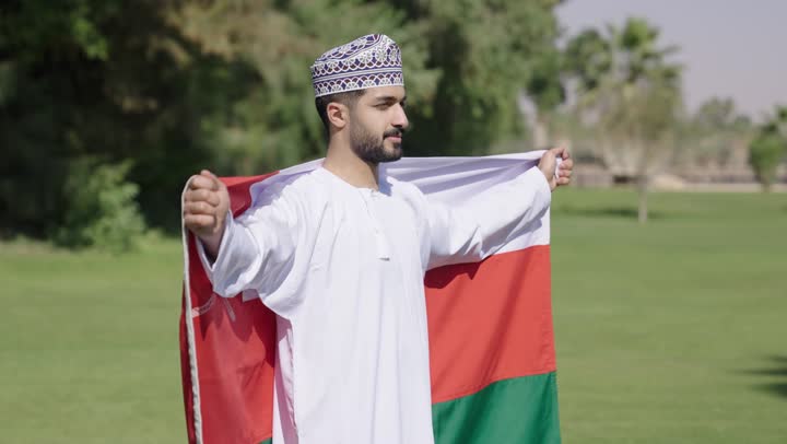 شاب عربي خليجي عماني يرتدي الدشداشة البيضاء و الكمة المطرزة يحمل العلم العماني  , الالتفاف مع النظر للكاميرا , الاعتزاز و الفخر بالوطن