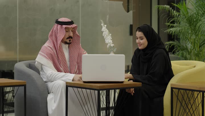 Teamwork and continuous collaboration to achieve the company's goals, the concept of cooperative management, a Gulf Arab Saudi man wearing a thobe and ghutrah sitting in front of a laptop with his colleague discussing work matters, completing daily office tasks.