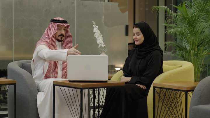 التكاتف والتفاعل بين فريق العمل لتحقيق أهداف الشركة والتغلب على المشكلات، مفهوم الإدارة المتعاونة، رجل عربي خليجي سعودي يرتدي الثوب و الشماغ يجلس امام الحاسب المحمول مع زميلتة يتناقش في أمور العمل، إنجاز المهام المكتبية اليومية