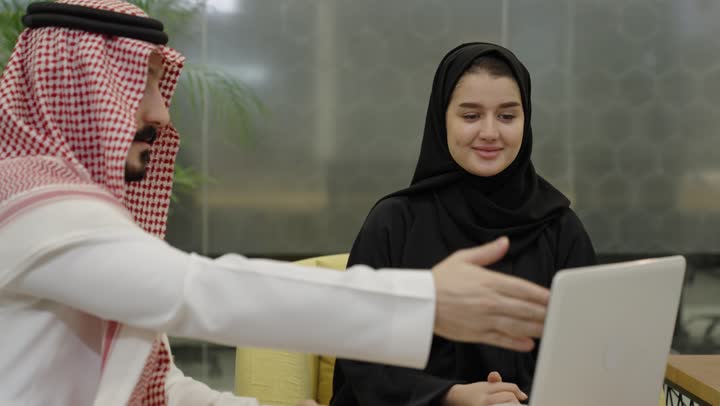تعزيز روح الوحدة والعمل الجماعي لتحقيق أهداف الشركة وحل المشكلات، مفهوم الإدارة المتعاونة، رجل عربي خليجي سعودي يرتدي الثوب و الشماغ يجلس امام الحاسب المحمول مع زميلتة يتناقش في أمور العمل، إنجاز المهام المكتبية اليومية