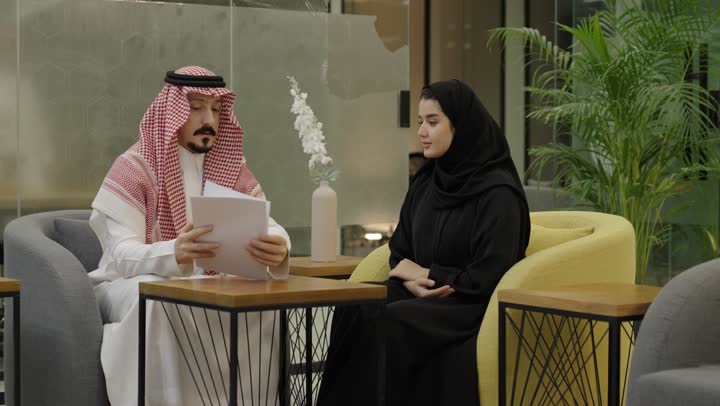 التعاون والتفاهم بين الفريق، شرح المهام المطلوبة، تنظيم وادارة جدول مفهوم العمل, تبادل الخبرات والاتفاقيات الناجحة, رجل عربي خليجي سعودي يرتدي الثوب الابيض و الشماغ يجلس برفقة زميلته يقوم بشرح العمل