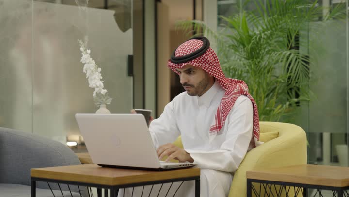 تطور الشركات السعودية، إتمام مهام العمل في المكتب، إدارة شؤون الأعمال وتنظيمها، احتساء مشروب القهوة، استخدام جهاز تقني حديث ومتطور، رجل عربي خليجي سعودي الثوب و الشماغ يستخدم الحاسوب، مهنة ووظيفة مكتبية، دمج العمل مع التقنية والتكنولوجيا