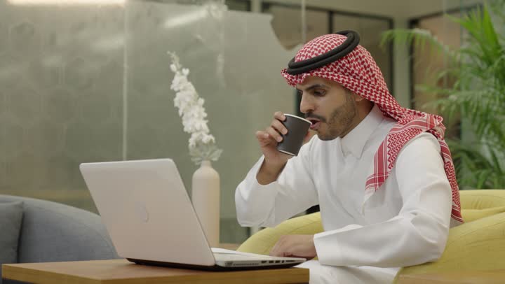 تطور الشركات السعودية، إتمام مهام العمل في المكتب، إدارة شؤون الأعمال وتنظيمها، احتساء مشروب القهوة، استخدام جهاز تقني حديث ومتطور، رجل عربي خليجي سعودي الثوب و الشماغ يستخدم الحاسوب، مهنة ووظيفة مكتبية، دمج العمل مع التقنية والتكنولوجيا