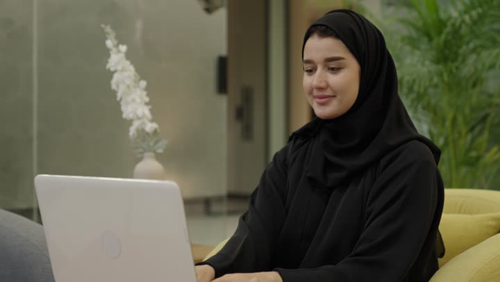 استخدام جهاز تقني حديث ومتطور، إتمام مهام العمل في المكتب، تطور الشركات السعودية، إدارة شؤون الأعمال وتنظيمها، احتساء مشروب القهوة،  امراه عربيه خليجية سعودية محجبة ترتدي العباءة تستخد الحاسوب المحمول، مهنة ووظيفة مكتبية، دمج العمل مع التقنية والتكنولوجيا