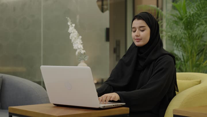 الوحدة والتفاعل الجماعي لتحقيق أهداف الشركة وحل المشكلات، مفهوم الإدارة المتعاونة، امراه عربيه خليجية سعودية ترتدي العباءة و الحجاب تجلس امام الحاسب المحمول تتناقش مع زميلها في أمور العمل، إنجاز المهام المكتبية اليومية