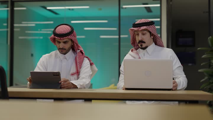 الاشارة الى شاشة الحاسوب، إنجاز الأعمال المطلوبة، الوحدة والعمل الجماعي، دمج الأعمال مع التقنية و التكنولوجيا، رجلان عربيان خليجيان سعوديان يرتديان الزي التقليدي يجلسان في المكتب يعملان على الحاسب المحمول وجهاز التابلت, تحقيق أهداف الشركة