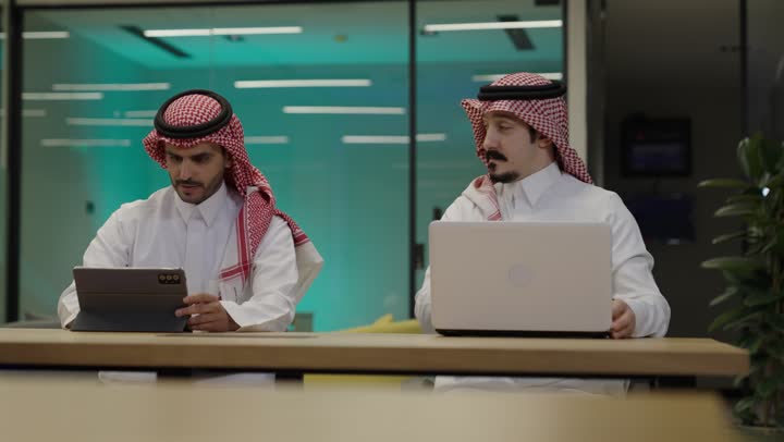 إنجاز الأعمال المطلوبة، الوحدة والعمل الجماعي، دمج الأعمال مع التقنية و التكنولوجيا، رجلان عربيان خليجيان سعوديان يرتديان الزي التقليدي يجلسان في المكتب يعملان على الحاسب المحمول وجهاز التابلت, تحقيق أهداف الشركة، الاشارة الى شاشة الحاسوب