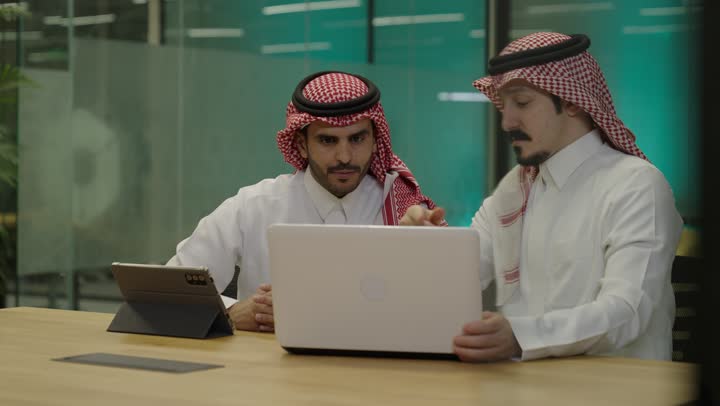 Unity and teamwork, integrating business with technology, accomplishing the required tasks, two Arab Gulf men from Saudi Arabia wearing traditional attire sitting in the office working on a laptop and a tablet, achieving the company's goals, pointing to the computer screen.