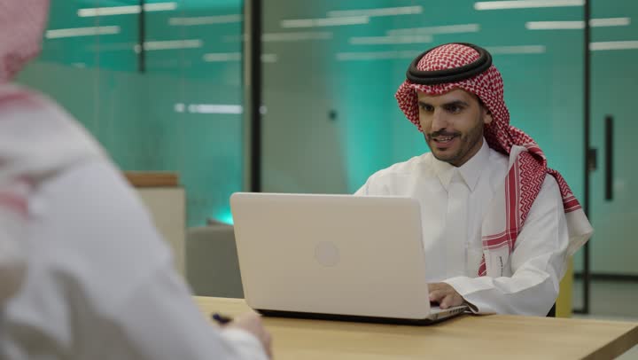 Making a video call over the internet, gestures of engagement and focus, using advanced modern technology in professional life, an office job and profession, an Arab Gulf Saudi man wearing traditional thobe and shemagh working on a laptop at the company headquarters.