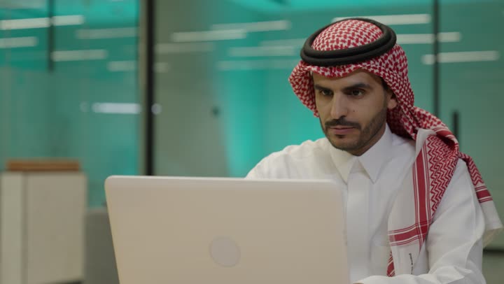 ايماءات الاندماج والتركيز، استخدام التقنية الحديثه المتطورة في الحياة العملية، مهنة ووظيفة مكتبية، رجل عربي خليجي سعودي يرتدي الثوب التقليدي والشماغ يقوم بالعمل على الحاسب المحمول في مقر الشركة
